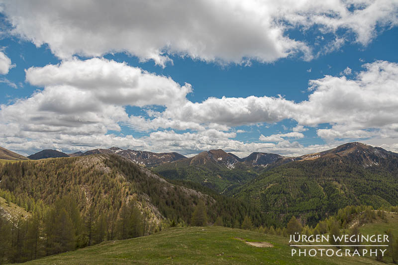 Weite Landschaft im alpinen Raum auf Berge und grüne Wiese. Blauer Himmel mit weissen Wolken. Sonnig. Nockalm Panoramastraße, Kärnten