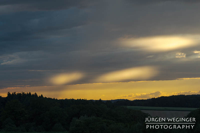Abendhimmel zum Sonnenuntergang. Landschaft als Silhouette abgebildet, dunkle Wolken mit einem orangen Band dazwischen.