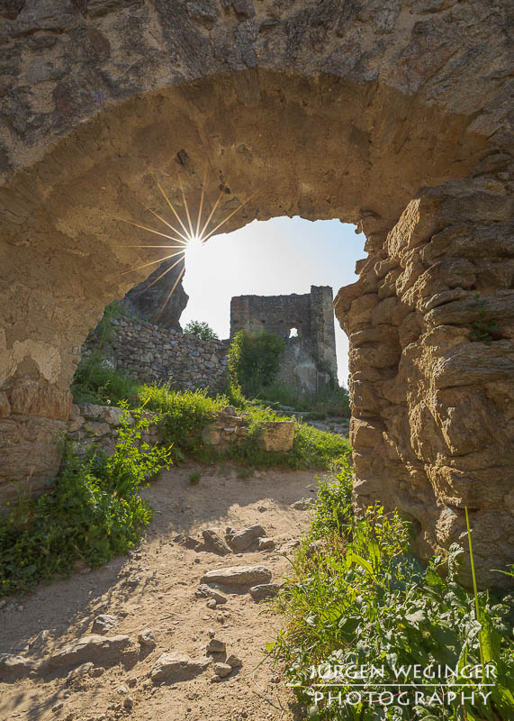 Pfad durch eine Burgruine mit verfallender Steinmauer und einem Torbogen mit Gegenlicht und Sonnenstern. Burgruine Dürnstein ,Wachau, Niederösterreich.