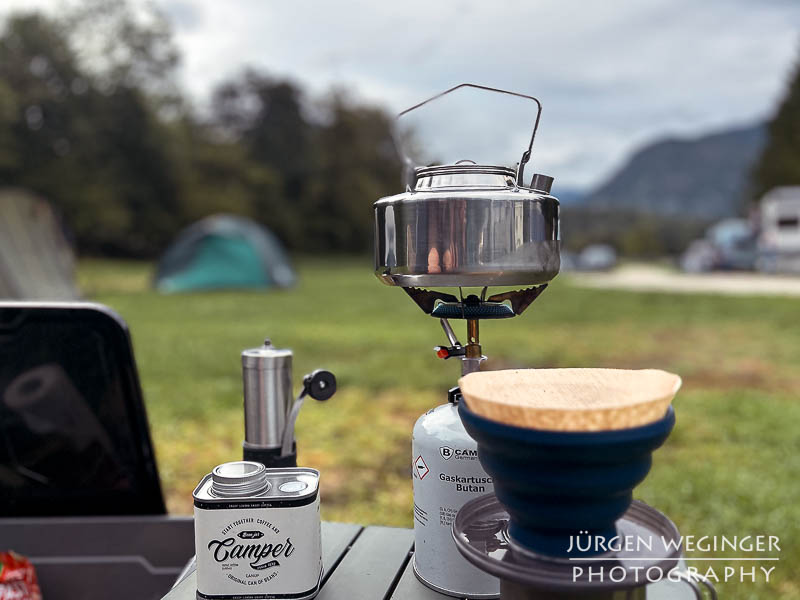 Camping Ausrüstung auf einen kleinen Tisch. Gaskocher mit kleinen Wasserkessel, Kaffeefilter und einer Metalldose mit der Aufschrift Camper